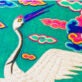 Vibrant embroidery depicting a white crane with a long neck and expansive wings, set against a turquoise background adorned with stylized pink, green, and blue clouds.